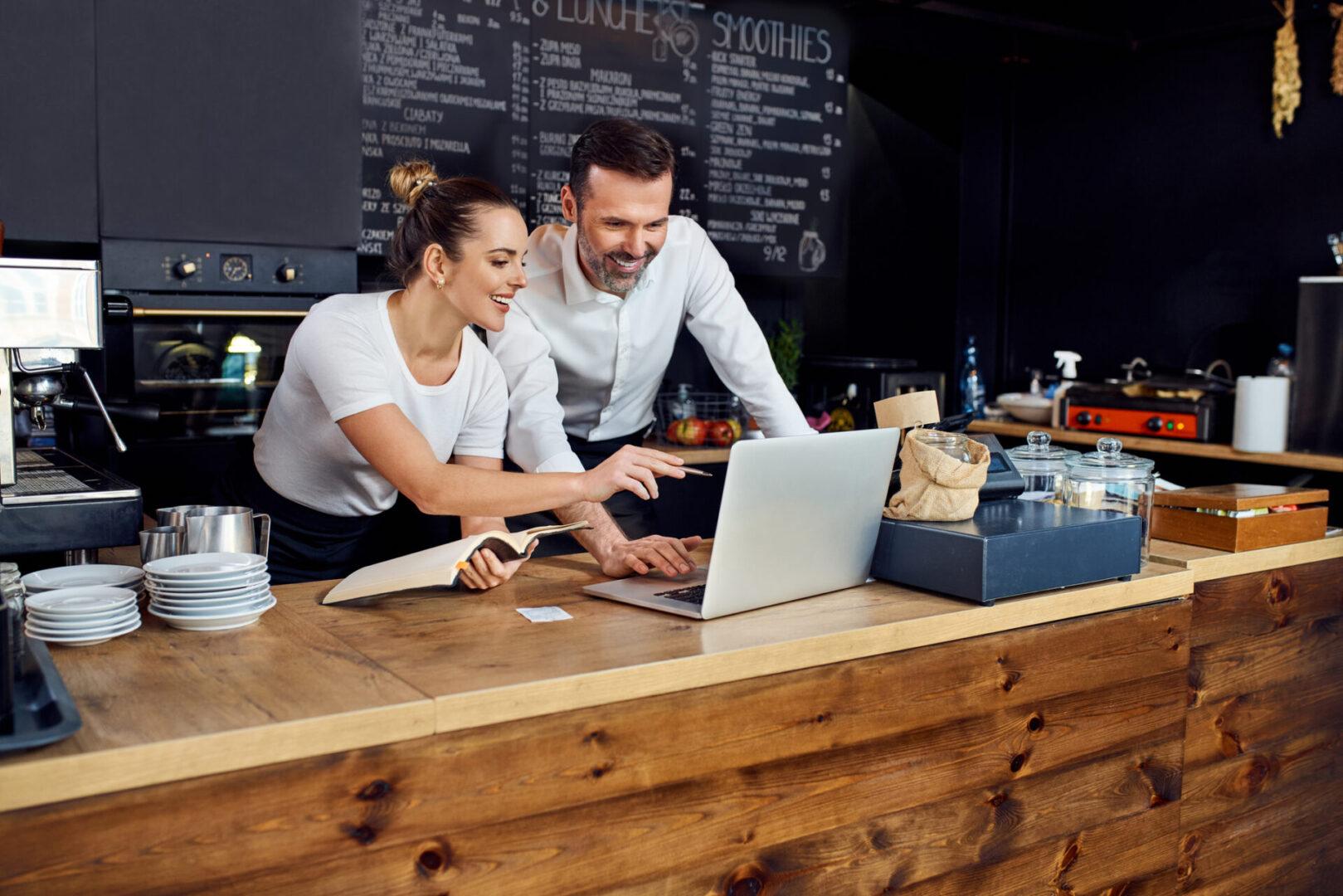 Two baristas collaborating over a laptop behind a coffee shop counter.