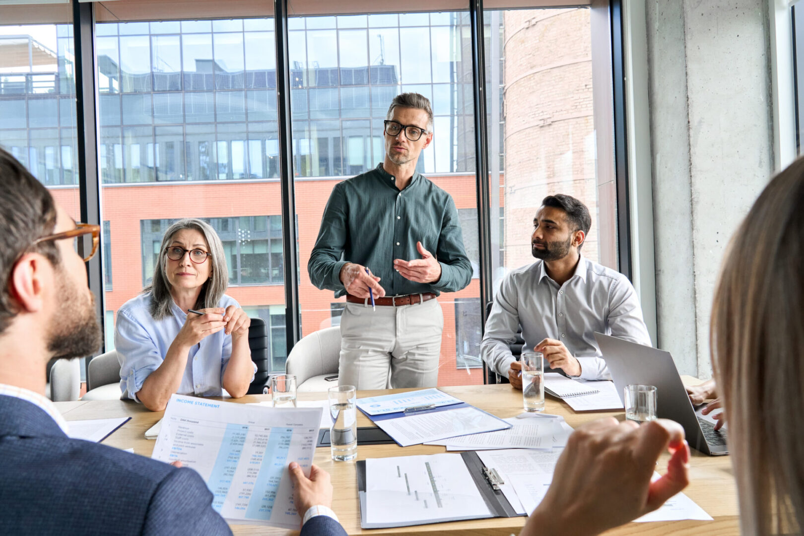 A man standing and speaking during a meeting with colleagues around a table.