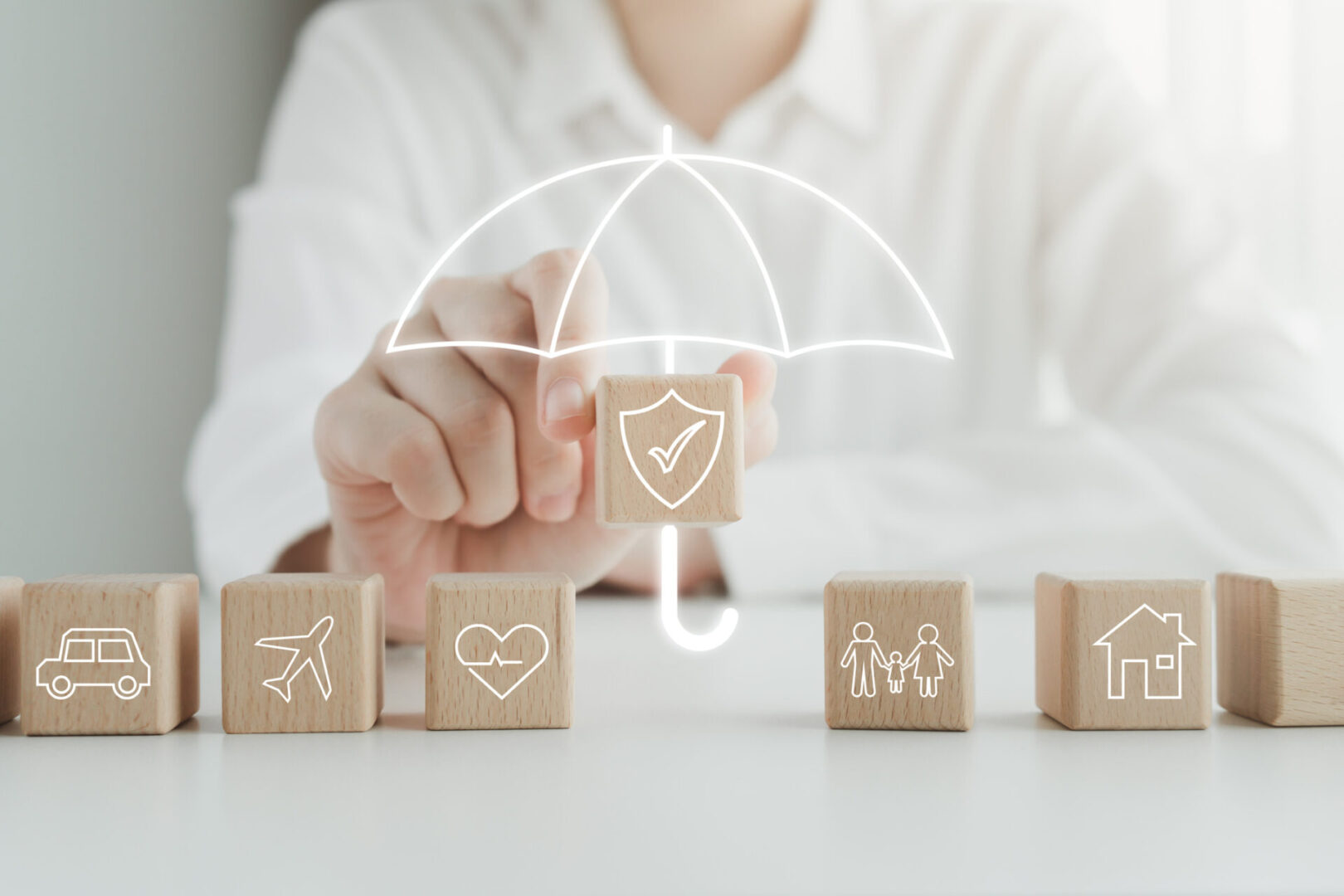 Hand protecting wooden blocks with insurance icons under a white umbrella.
