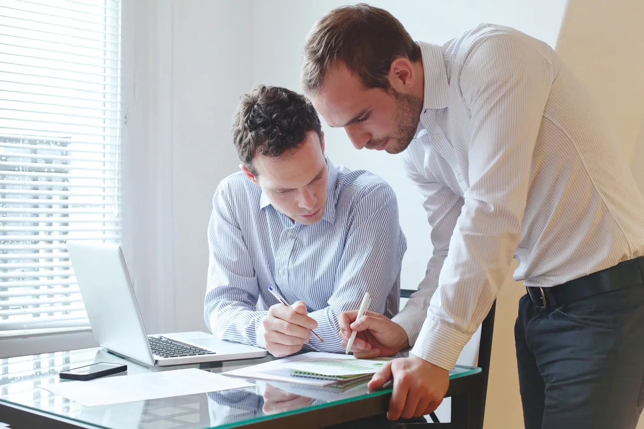 Two men collaborating on a project at a desk with documents and a laptop.
